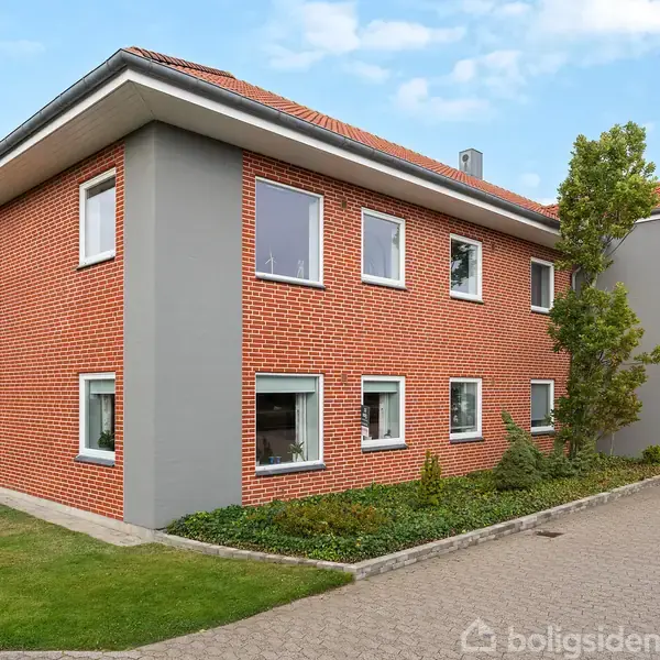 En beige sofa står i en stue med trægulv, flankeret af et sofabord. En spiseplads med bord og stole ses i baggrunden ved siden af et stort skab med dekorative genstande.