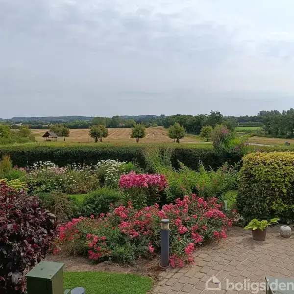 En farverig have med blomstrende buske og planter pryder forgrunden. I baggrunden ses marker og spredte træer på en rolig, landlig horisont under en skyet himmel.