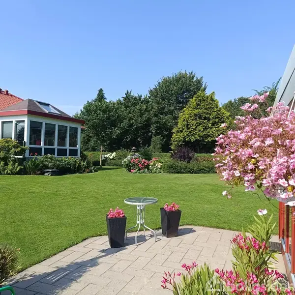 En velplejet have med en terrasse med bord, to blomsterkrukker, og lyserøde blomster hængende. I baggrunden ses et glashus med rødt tag og grønne træer.