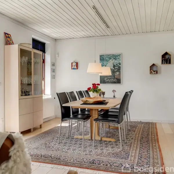 Et spisebord med sorte stole står på et orientalsk tæppe i en lys spisestue. En glasvitrineskab står ved vinduet, og kunst pryder væggene.