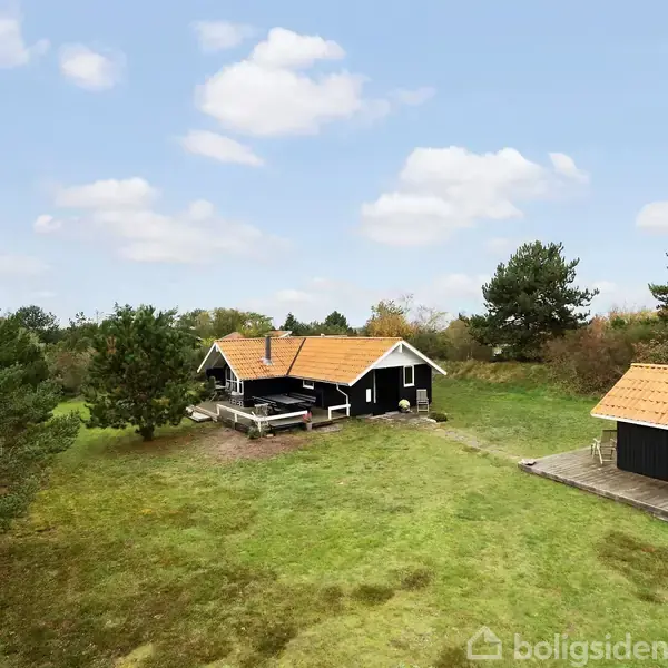 To sorte huse med røde tegltage befinder sig på en grøn græsplæne omgivet af træer under en skyet himmel.
