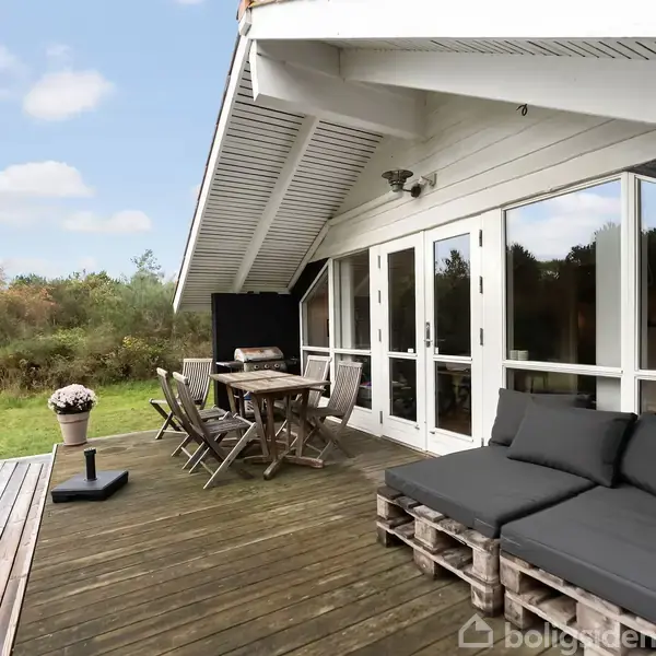 Træterrasse med sofagruppe af paller og hynder, og spisebord med stole, vender ud mod græsplæne og buske i baggrunden. Hvidt hus med store glasdøre ses ved siden.