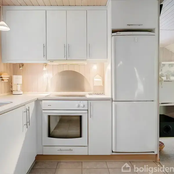 Hvidt køkken med indbyggede skabe og apparater. Komfur og ovn er centreret under skabe. Til højre ses et køleskab. Vindue med lysindfald til venstre. Flisegulv.