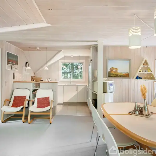 Stue med trævægge og loft. To stole med røde puder står ved siden af et moderne køkken. Et spisebord med stole er i forgrunden. Maleri og hylder på væggen.