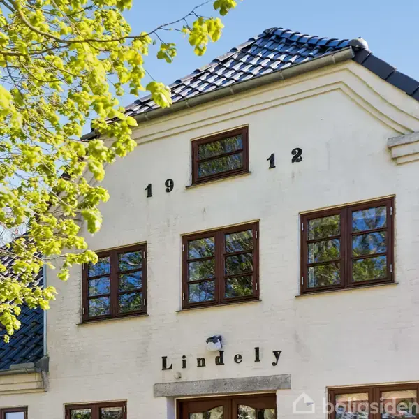 Hvid bygning med blå tegltag og trævinduer viser årstallet "1912" og navnet "Lindely" på facaden. Grønne træer omgiver huset.