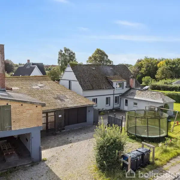 Et hus med en tilhørende bygning, hvor en skorsten findes på bygningen. En trampolin står i haven. Omgivet af en vej og andre huse i et grønt område.