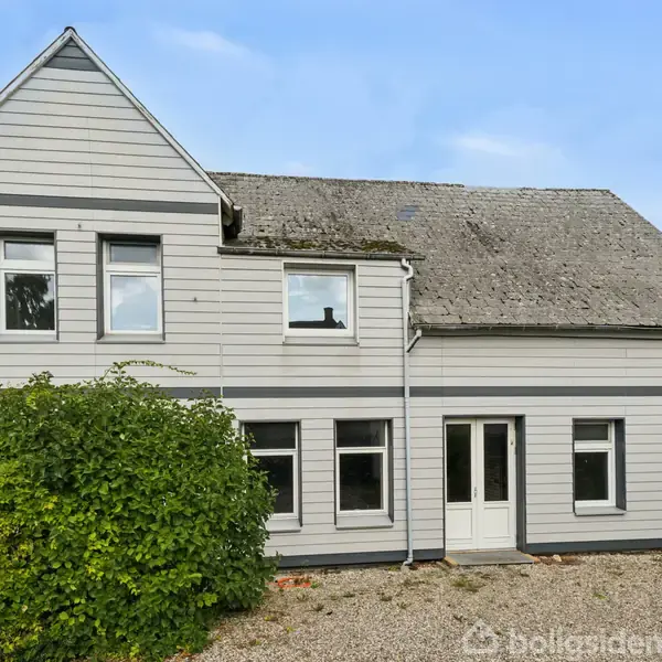 Et gråt, to-etagers hus står stille i en grusbelagt gårdsplads, omgivet af grønne buske og træer under en blå himmel.