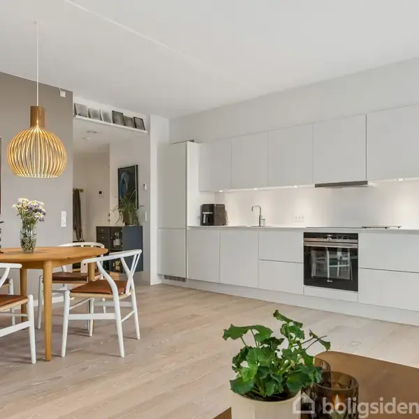 Et moderne køkken med hvide skabe og en integreret ovn strækker sig langs væggen. Rundt spisebord med stole er oplyst af en pendellampe. En maleri hænger på den grå væg.