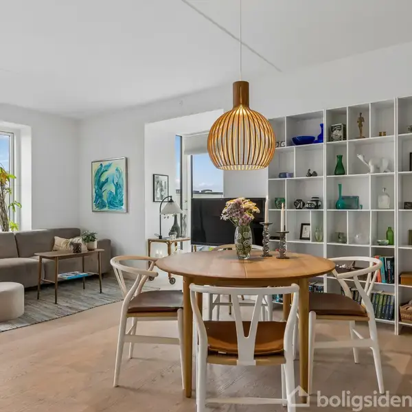Rundt spisebord med stole og blomster i vase; en stue med sofa og dekorerede hylder i baggrunden; moderne, lyst rum med kunstværker og grønne planter.