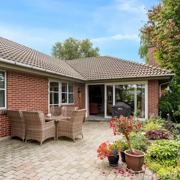 Brun murstenshus med terrasse. Rattanmøbler er placeret på fliserne. Omkring terrassen er blomsterkrukker og grønne træer, hvilket skaber en hyggelig haveatmosfære under en klar blå himmel.