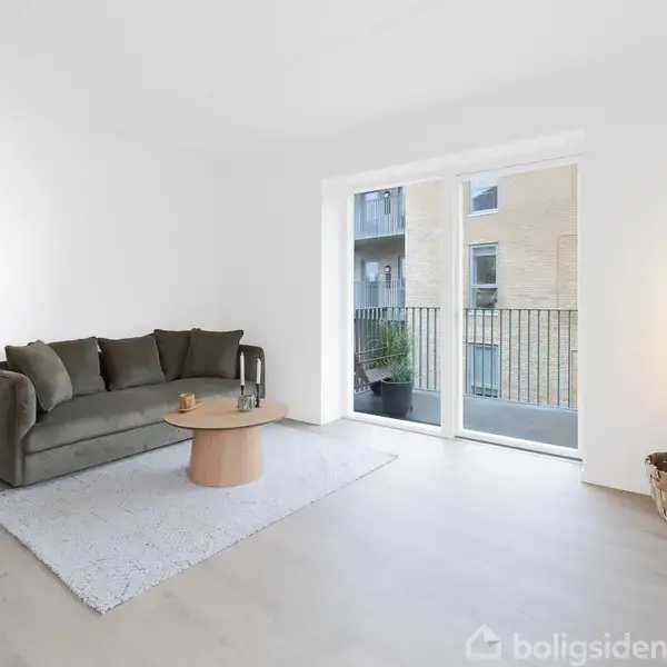 En grøn sofa står på et lyst tæppe ved siden af et rundt træbord i en minimalistisk stue med en stor vinduesåbning til en altan.