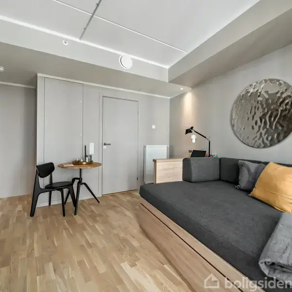 Sofa med grå puder og gult pude (objekt) står i et minimalistisk værelse (kontekst) med trælamingulv, sort stol, bord med stearinlys og moderne kunst på væggen (handling).