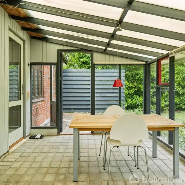 Et træbord med to hvide stole står under et overdækket terrasseområde med glaspaneler. En rød lampe hænger over bordet. Døren fører udenfor til en have med grøn beplantning.