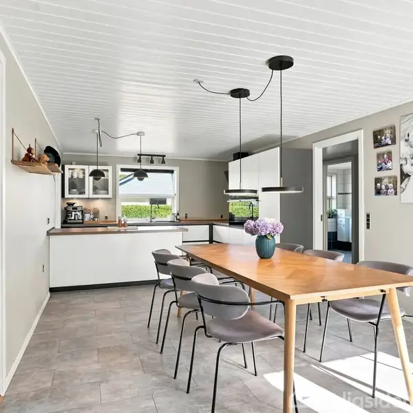Træbord med stole står i lys spiseplads. Køkken ses i baggrunden med hvidt interiør. På væggen hænger et stort billede med familiefotos. Blomster i vase pryder bordet.