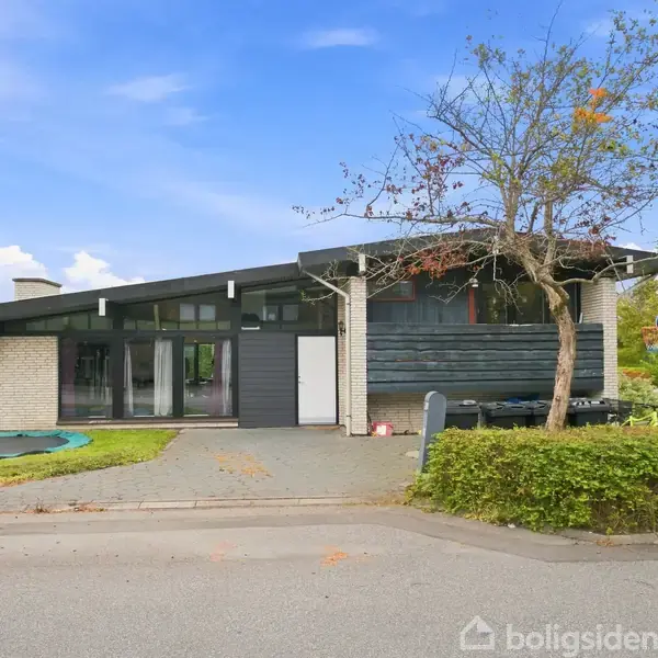 Et moderne hus står stille i en boligkvarter med hvid murstensfacade og sort træværk. En trampolin er indlejret i græsset i forhaven, omgivet af hække og træer.