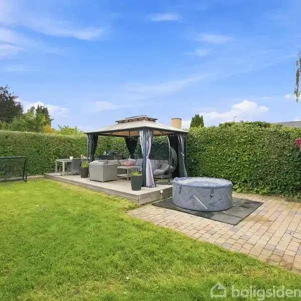 Pavillon med sofaer og et bord står på en terrasse. Ved siden af er et oppusteligt spabad. Græsplæne og hække omgiver området, basketballkurv er monteret på et skur.