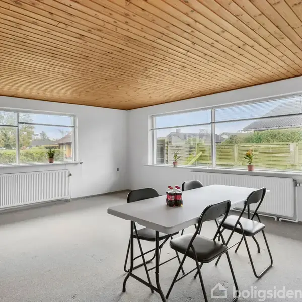 Et bord med tre stole står i et lyst rum med træloft. Der er store vinduer med udsigt til haven, og tre planter står på radiatorerne. Flasker står på bordet.