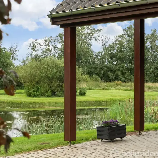Terrasse med træstolper står foran en rolig sø omgivet af græs og træer, hvilket skaber en fredfyldt, naturskøn scene i en have eller park.