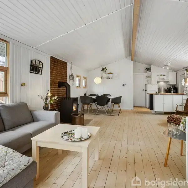 Stue med gråt sofaarrangement ved et hvidt sofabord. Pejs mod en murstensvæg, spisebord med stole, og et åbent køkken i baggrunden. Trægulv og hvidmalet loft.