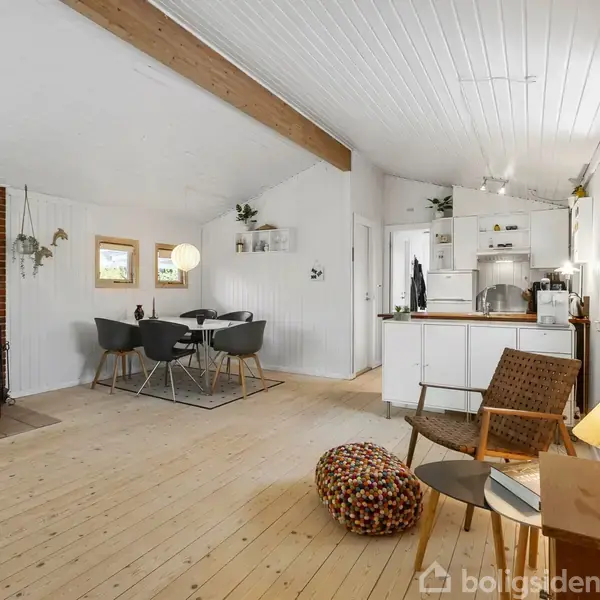 Brændeovn står til venstre og opvarmer stuen med plant gulv. Træloft og hvide vægge skaber en lys atmosfære. Til højre er spiseområde og køkken med moderne møbler.