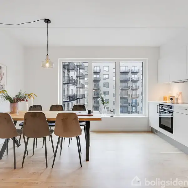 Et lyst køkken-alrum med et træbord omgivet af seks stole. Et stort vindue viser en udsigt over en moderne bygning. Køkkenet har hvide skabe og en sort ovn.