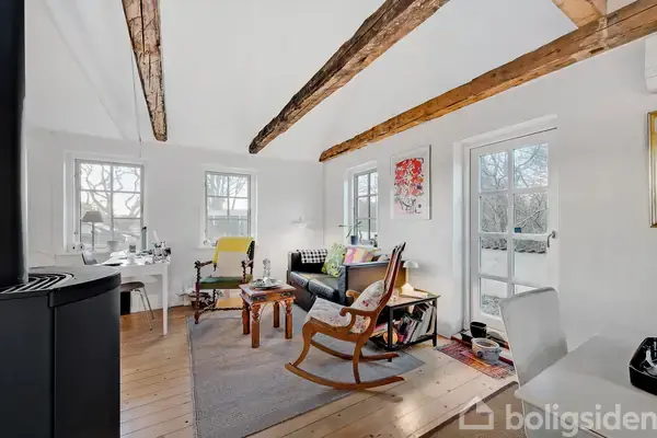 Trærstol gynger i lys stue med synlige træbjælker, farverig kunst og møbler. Store, lyse vinduer ud mod natur, gulvtæppe på trægulv, og brændeovn i hjørnet.