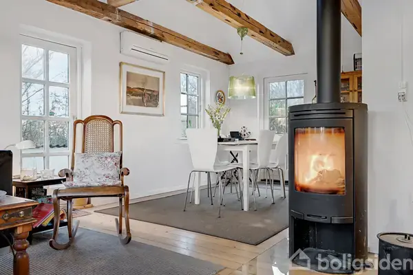 Brændeovn med flammer opvarmer en hyggelig stue. I baggrunden står et hvidt spisebord med stole under synlige loftsbjælker. Der er vinduer med udsigt til en frodig have.