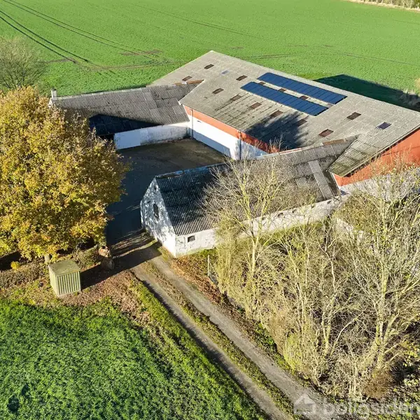 En gårdbygning med stald og redskabsskur, set i fugleperspektiv, omkranset af træer og åbne marker. Vej fører op til gården, mens solpaneler ses på taget.