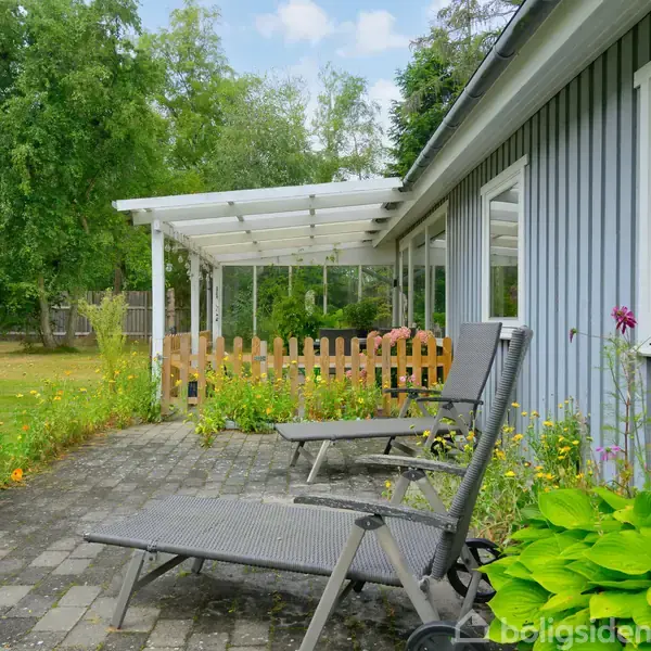 To liggestole står på en terrasse ved siden af et gråt hus med store vinduer. Bag terrassen er en lille have med græs, blomster og store træer under en blå himmel.