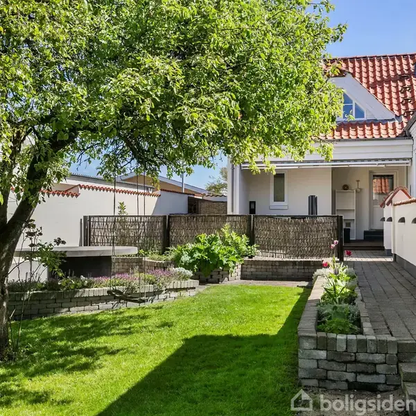 Hus med hvid facade og rødt tegltag står i en solrig have. Træ langt forrest i billedet. Flisebelagt gang leder til indgang, omkranset af grønne planter og muret blomsterbed.