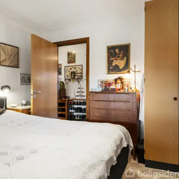 En seng med hvidt sengetæppe står i et soveværelse med træmøbler. En dør står delvist åben, og væggene har forskellige kunstværker. En lampe lyser ved sengen.