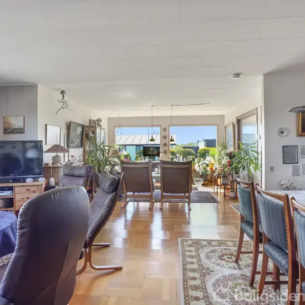 Stue med spisebord, sofaer og lænestole. TV på skænk, blå dug på bordet, lysekrone og malerier på væggen. Store vinduer åbner op til en terrasse med udsigt.