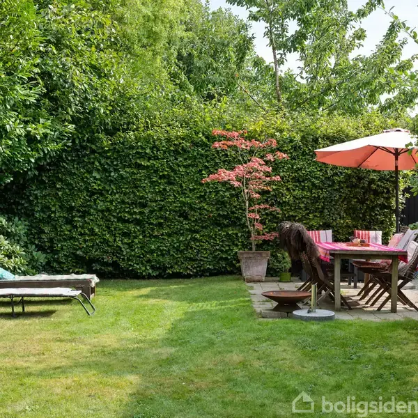 En solseng står på en græsplæne. Et bord med stole er dækket med en lyserød dug og parasol. Omkring er der høje grønne buske og træer.