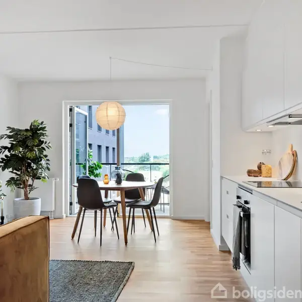 Et moderne køkken i åbent plan er forbundet med en lys spisestue. Et spisebord med fire stole står foran en stor, åbnet dør mod en balkon. En loftslampe hænger over bordet.