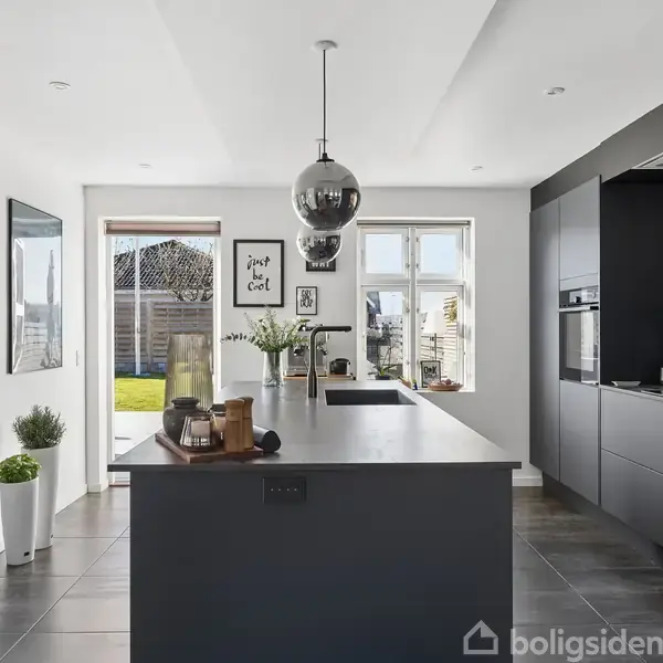 Køkken med en stor mørkegrå ø i midten, blomster i vaser og hængende lampe. Moderne skabe i baggrunden. Kunst på væggene inkluderer tekst: "Just be cool". Udsigt til have.