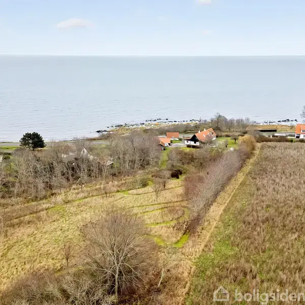 Spredte huse ligger tæt ved kysten, mens en mark strækker sig i forgrunden omgivet af træer. Havet fylder horisonten under en klar himmel med få skyer.