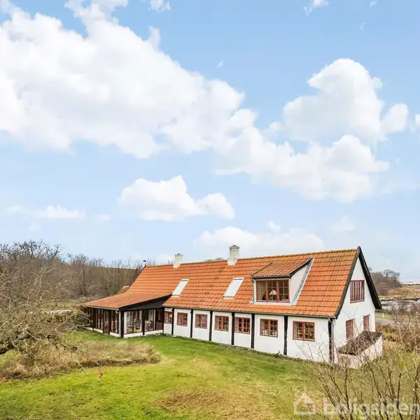 Et hvidt, stråtækket hus med rødt tag ligger i en åben, grøn have med træer, under en blå himmel med spredte skyer.