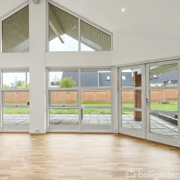 Rummet har trægulv og store vinduespartier, der åbner op mod en terrasse og en have med hæk. Lyse vægge og højt, skråt loft skaber en rummelig atmosfære.
