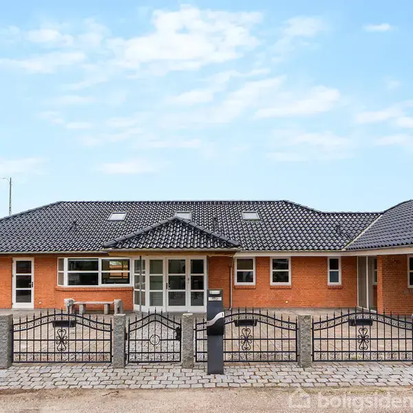 En enfamiliesvilla med røde mursten og sort tegltag står bag sort metalhegn. Døren og vinduerne er hvide. Huset er omkranset af blå himmel og nogle træer.