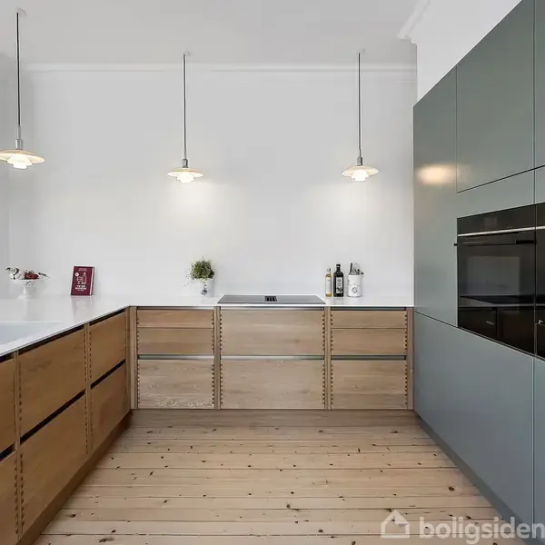 Et moderne køkken med trægulv og minimalistikke træskabe. Tre hængende lamper oplyser rummet. Der er en vask og en håndvask. Dekorationer inkluderer planter, bøger og en kraniets vægdekoration.