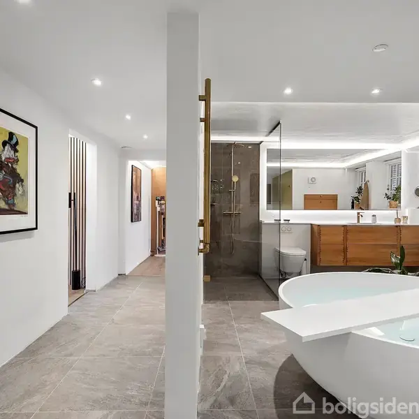 Et badeværelse med et fritstående badekar fyldt med vand og et glas vin på kanten, bagved en stor spejl- og vaskenhed. Hvide vægge og marmorgulv skaber en luksuriøs atmosfære.