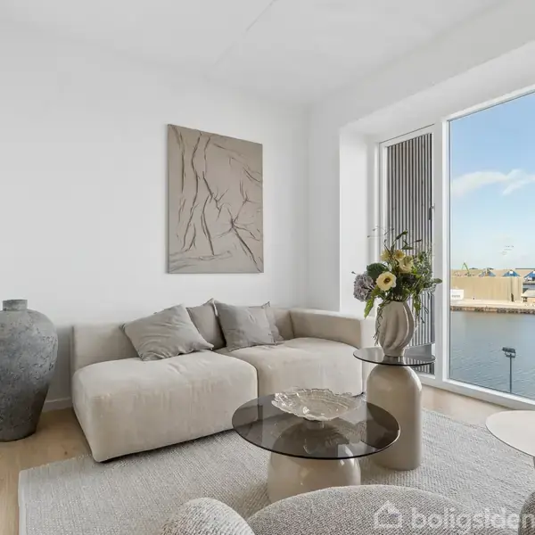 Sofa med grå puder står i en moderne stue med lyst trægulv. Et stort vindue med udsigt til vand og himmel. En vase med blomster står på et glasbord.