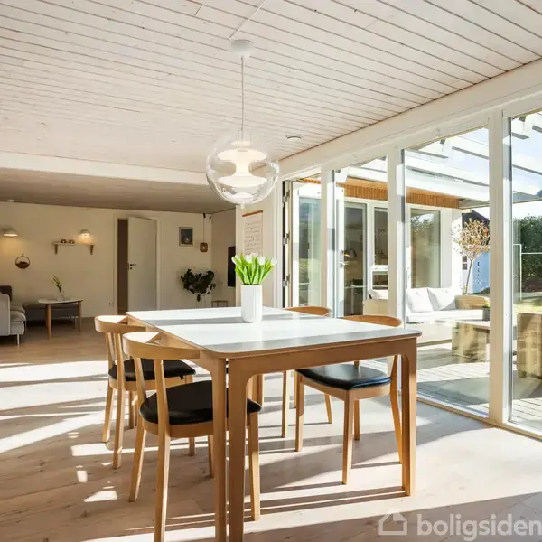 Et lyst spisebord med fire stole står i et rummeligt og solrigt rum. Store vinduer og en glasdør åbner op til en terrasse med en sofa. En vase med blomster står på bordet.