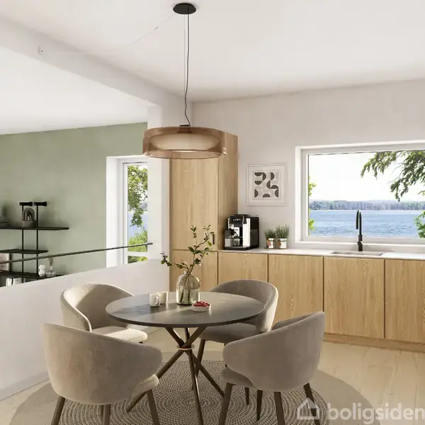Rundt spisebord omkranset af fire beige stole står i et lyst køkken med træskabe. Et vindue med udsigt over vandet giver naturligt lys.