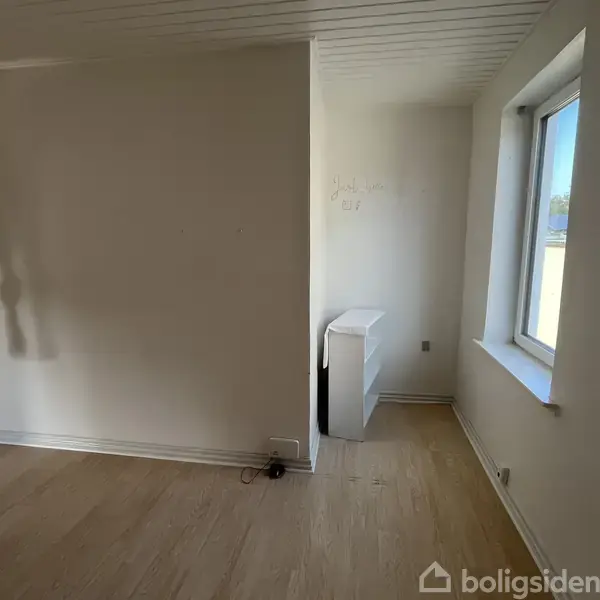 Et rum med en hvid væg og trægulv, tilstødende indhak med vindue. På væggen står teksten "Hang loose, summer is close." Rummet er lyst og tomt.