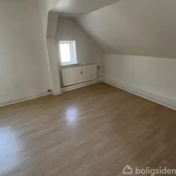 Et tomt værelse med lyse trægulve. Et vindue og radiator på bagvæggen giver indtryk af naturligt lys. Skråloftet skaber en hyggelig atmosfære.