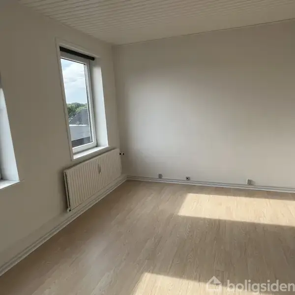 Et tomt rum med lyse trægulve, to vinduer med sorte rullegardiner og en radiator mellem dem. Udsigt til himmel og omkringliggende bygninger udenfor.