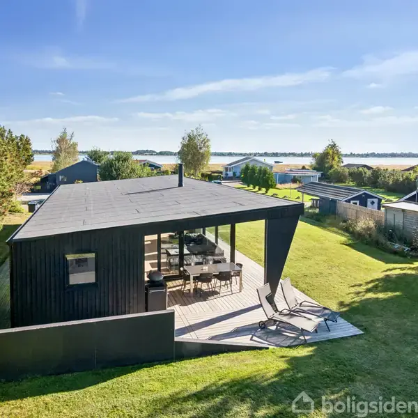 Sort moderne hus med terrasse, står stille på en grøn græsplæne. To liggestole er på terrassen, mens andre små huse og en sø kan ses i baggrunden under en blå himmel.
