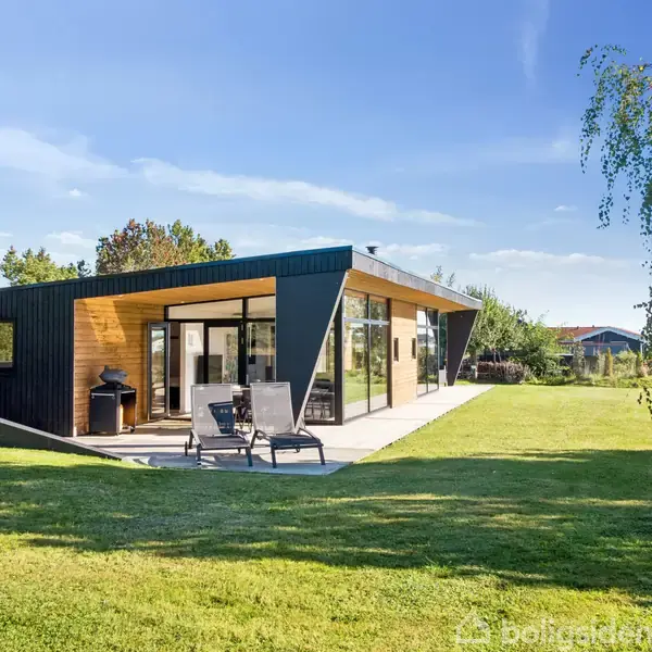 Et moderne, fladt taghus står i en grøn have med græsplæne. To liggestole er placeret på terrasseområdet ved huset, omgivet af træer og med klar blå himmel i baggrunden.