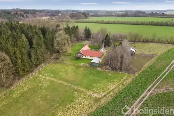 Hus med rød tag omgivet af træer og græsarealer, set fra luften. Beliggende i landligt område med marker og skov i baggrunden.